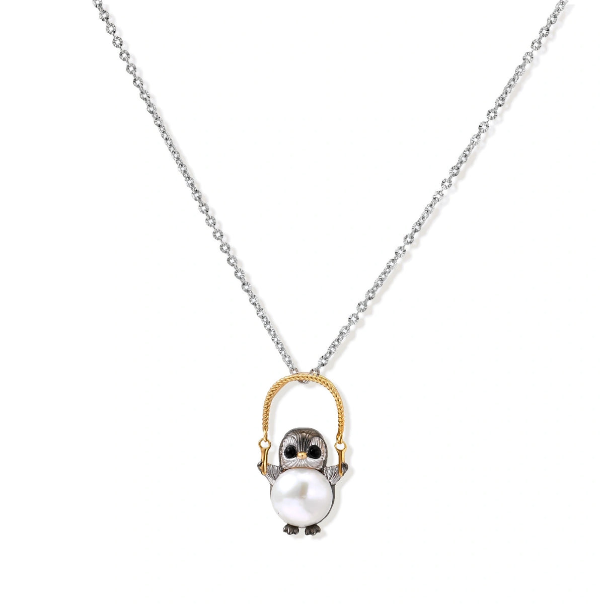 Whimsical penguin pendant holding a white pearl on a gold swing, rhodium-plated silver chain on cream background.