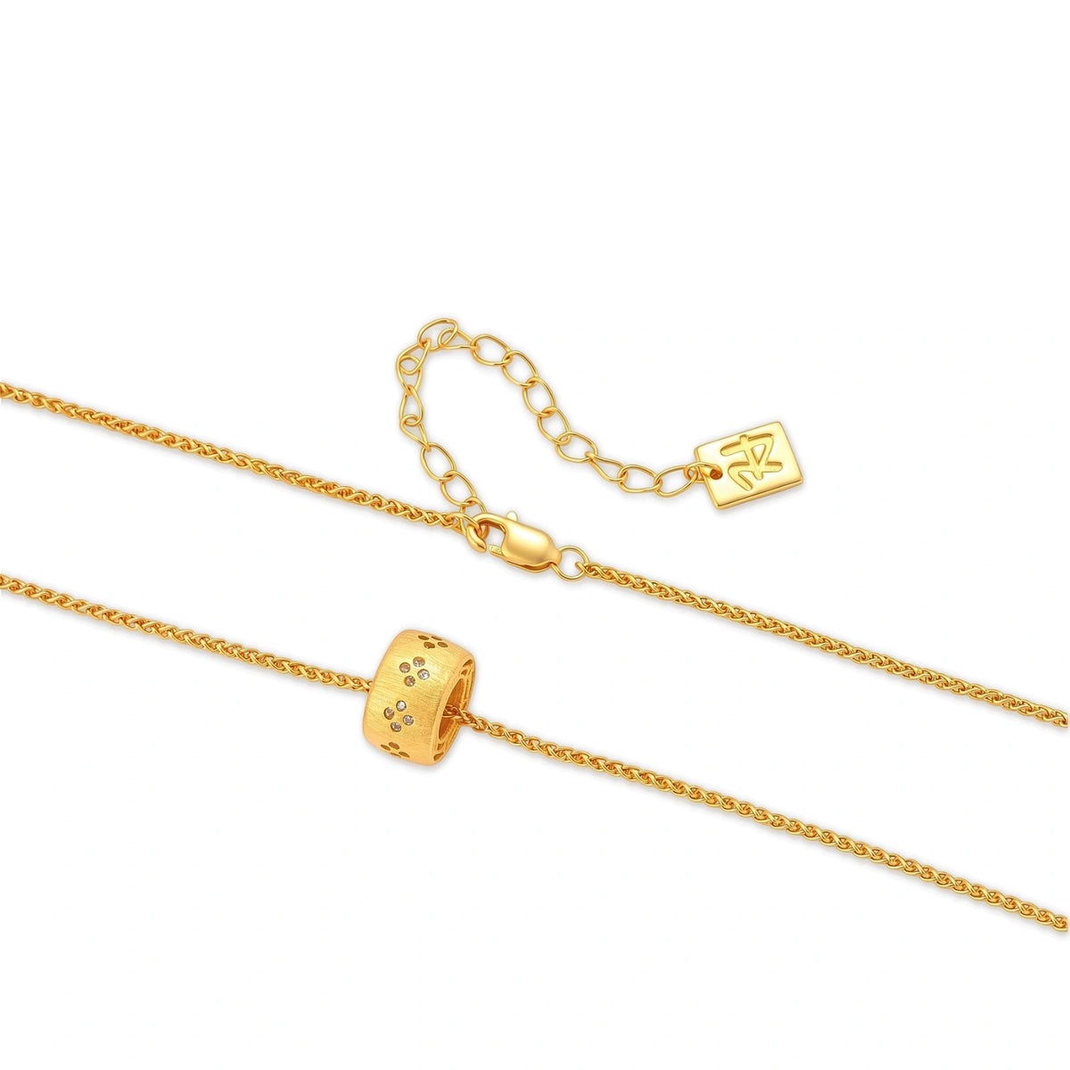 Gold-plated cylindrical ring pendant with brushed finish and scattered crystal accents on a wheat-link chain, cream background.